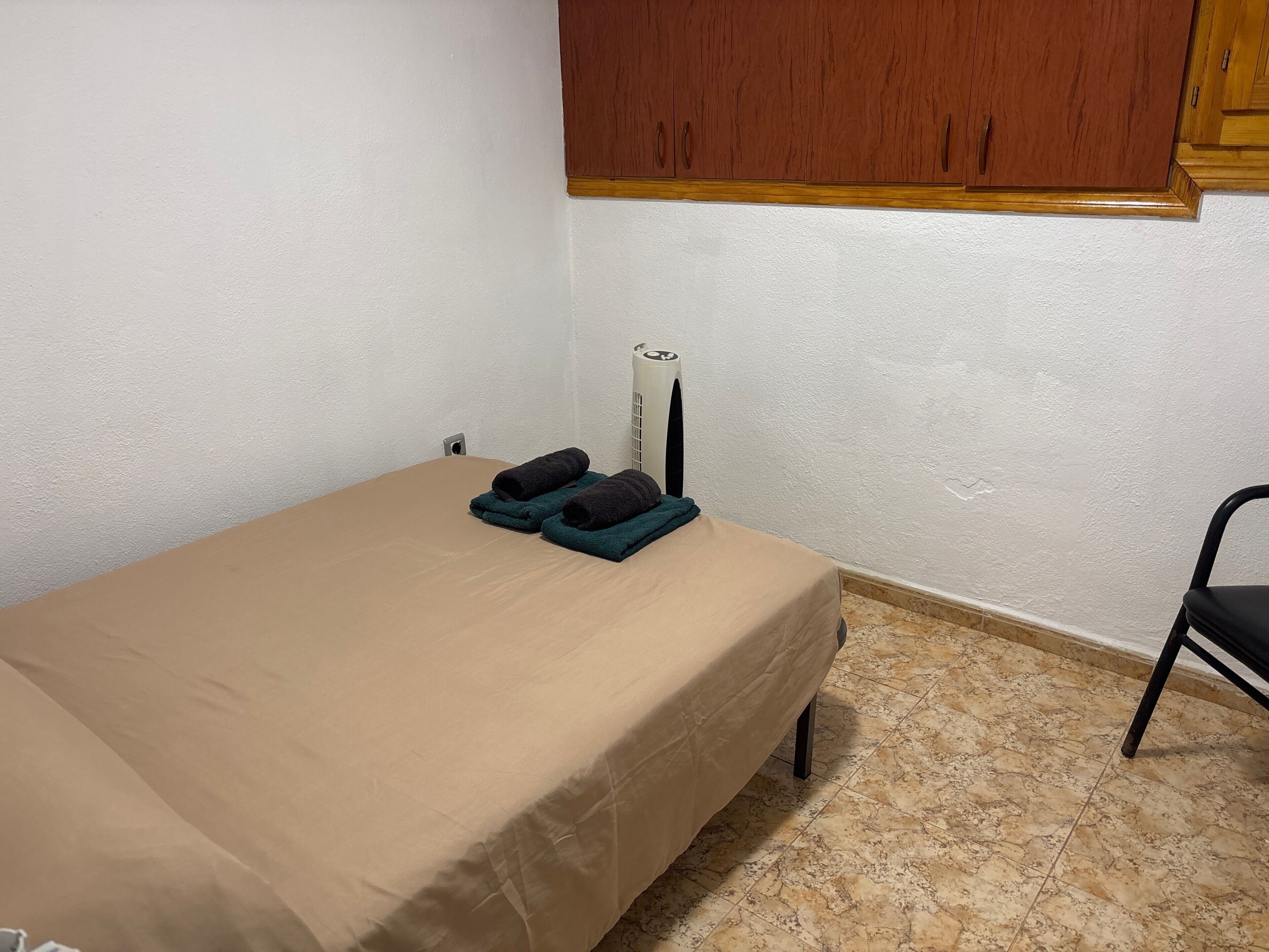 Una habitación pequeña con cama individual, colchón color beige, una silla negra y un armario de madera sobre la cama.