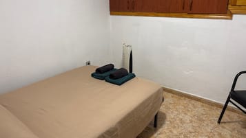 Una habitación pequeña con cama individual, colchón color beige, una silla negra y un armario de madera sobre la cama.
