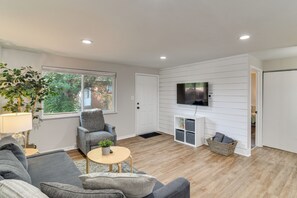 A modern living room with a grey sofa, a small round table, a television mounted on the wall, and a large window with a view of greenery outside. - 12 Mi to Red Rocks, Dtwn Denver! Townhome w/ Patio (Arvada)