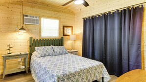 Una habitació de fusta amb un llit, una tauleta de nit, una làmpada, una finestra amb persianes i una cortina.