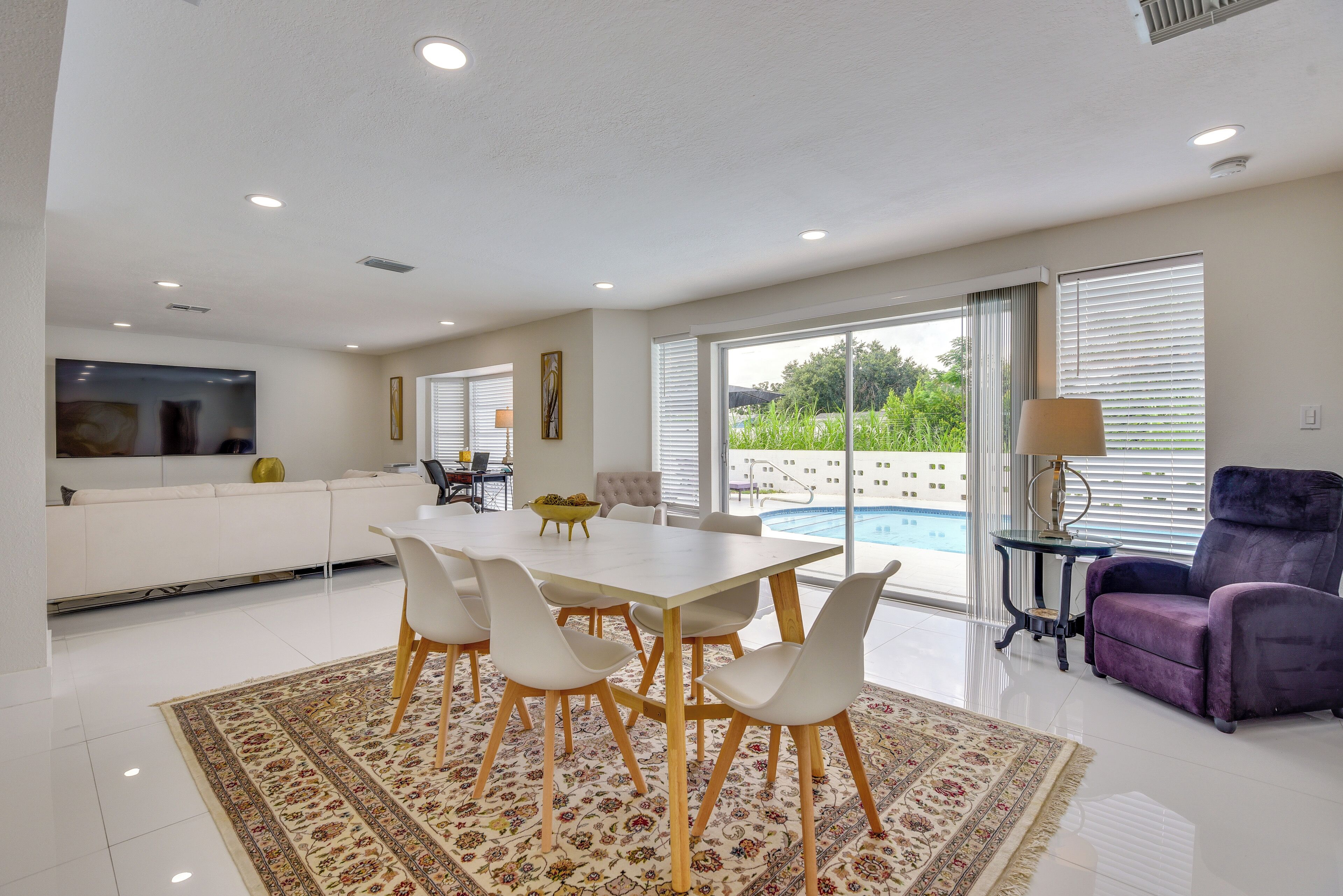 A modern living room with a dining table, white chairs, a purple armchair, and a view of a swimming pool.