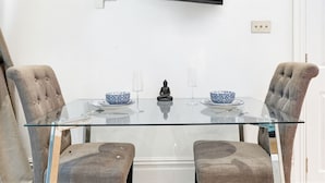 A modern dining area with a glass table, two cushioned chairs, a Buddha statue, and a television mounted on the wall. - Pet Friendly Studio in Fulham, Sleeps 2 (London)