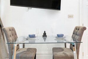Ein zeitgemäßer Essbereich mit einem Glastisch, zwei gepolsterten Stühlen, einer Buddha-Statue und einem an der Wand montierten Fernseher.