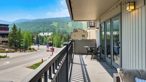 Un balcone con vista su una strada e montagne, un tavolo con sedie e un edificio con ampie finestre.