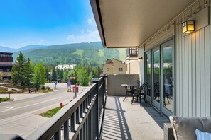 Ein Balkon mit Blick auf eine Straße und Berge, ein Tisch mit Stühlen sowie ein Gebäude mit großen Fenstern.