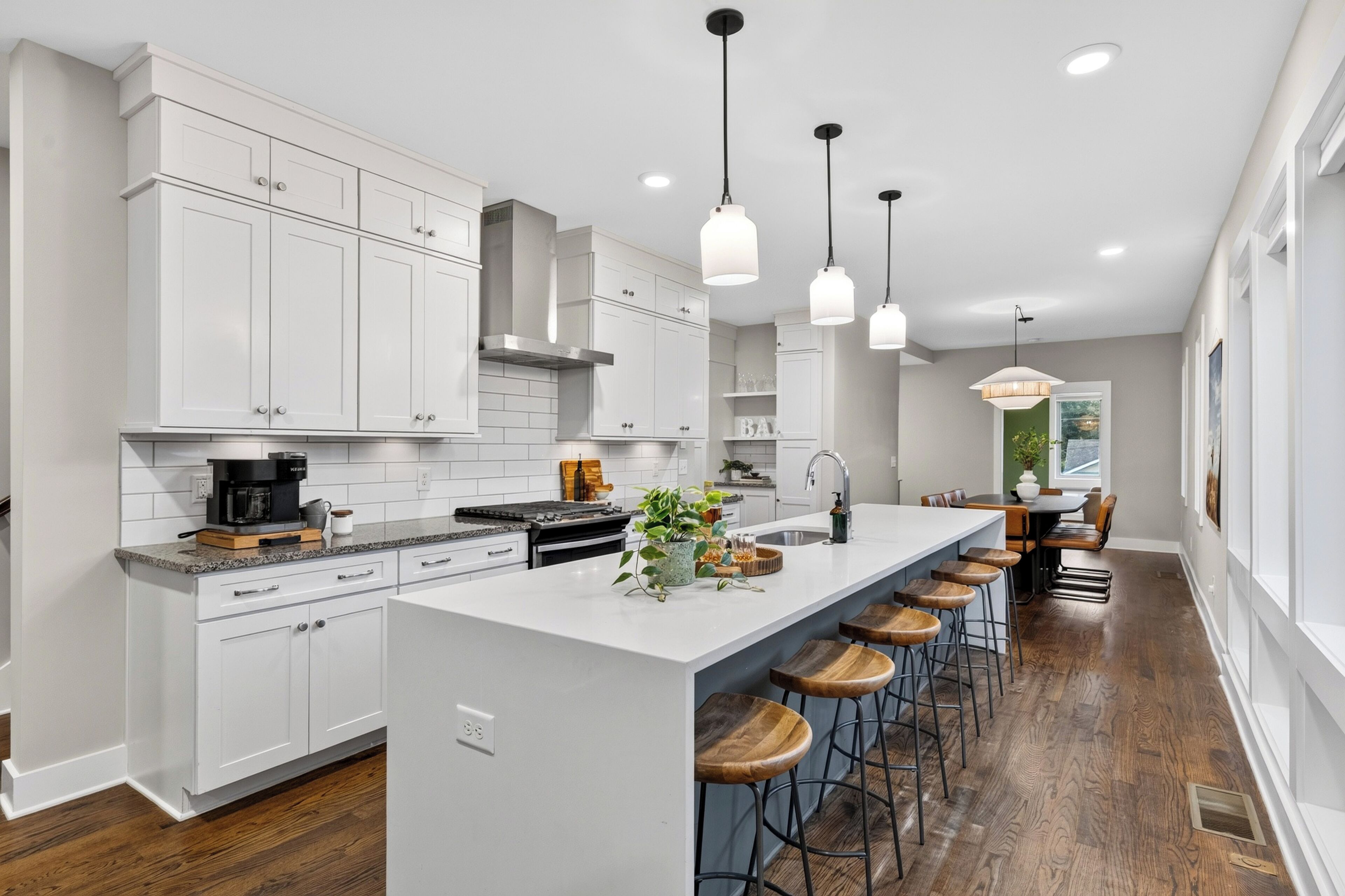 Une cuisine moderne avec des meubles blancs, un îlot central doté de tabourets, un éclairage suspendu et une vue sur la salle à manger.