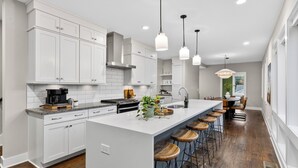 A modern kitchen with white cabinetry, a central island with stools, pendant lighting, and a view to the dining area. - Hot Tub + Rooftop Lounge + Game Room | Buchanan Arts | The Mod Buchanan (Nashville)