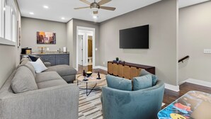 A living room with a gray sofa, a blue armchair, a flat-screen TV, a ceiling fan, and a kitchen area in the background. - Hot Tub + Rooftop Lounge + Game Room | Buchanan Arts | The Mod Buchanan (Nashville)