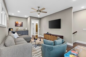 Ein Wohnzimmer mit grauem Sofa, blauem Sessel, Flachbildfernseher, Deckenventilator und einer Küche im Hintergrund.