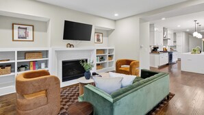A modern living room with a fireplace, a television, and a kitchen area with white cabinets and a central island. - Hot Tub + Rooftop Lounge + Game Room | Buchanan Arts | The Mod Buchanan (Nashville)