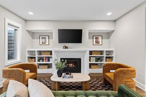 A living room with a fireplace, two armchairs, a coffee table, and a television mounted above the fireplace. - Hot Tub + Rooftop Lounge + Game Room | Buchanan Arts | The Mod Buchanan (Nashville)