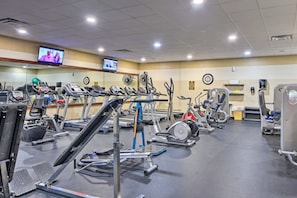A gym with various exercise machines, a television displaying a program, and a clock on the wall. - Screened Porch + Home Office: Modern Ellijay Cabin (Ellijay)