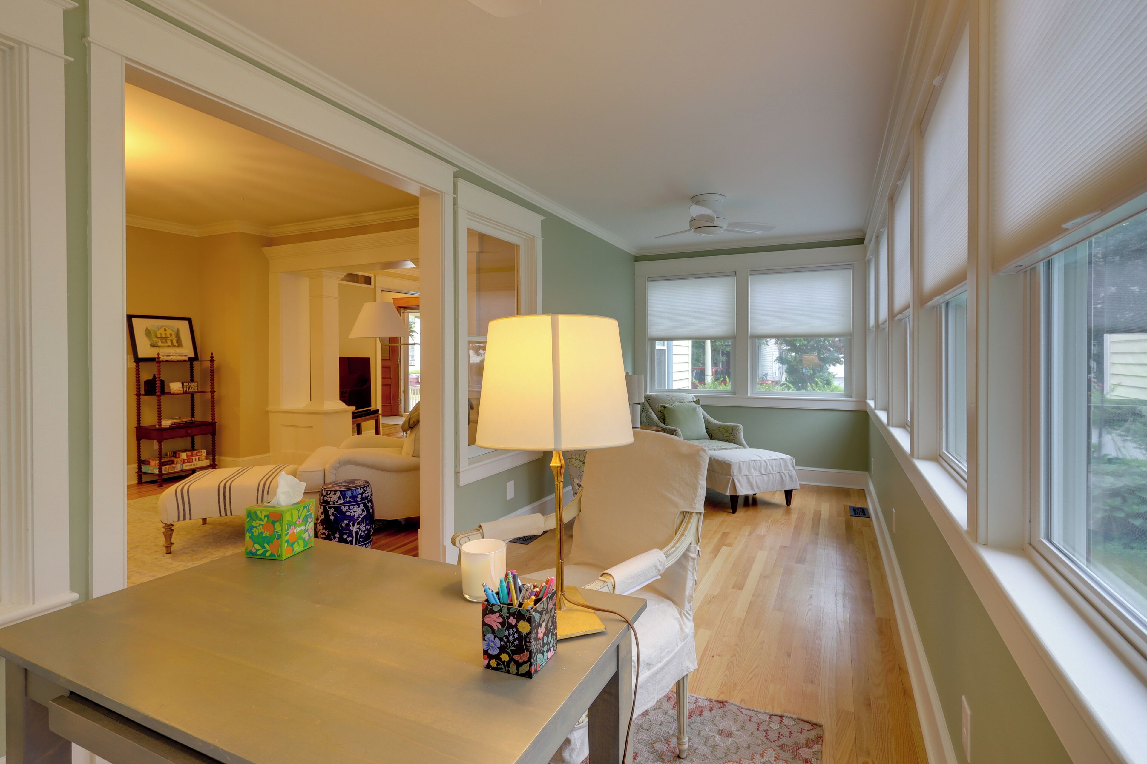 A room with a wooden floor, a table with a lamp, a chair, and a window with blinds.