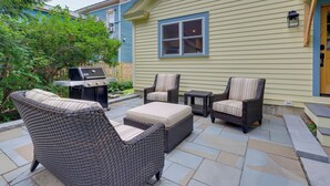 Een terras met rieten meubilair, een barbecue en een huis met blauwe bekleding.