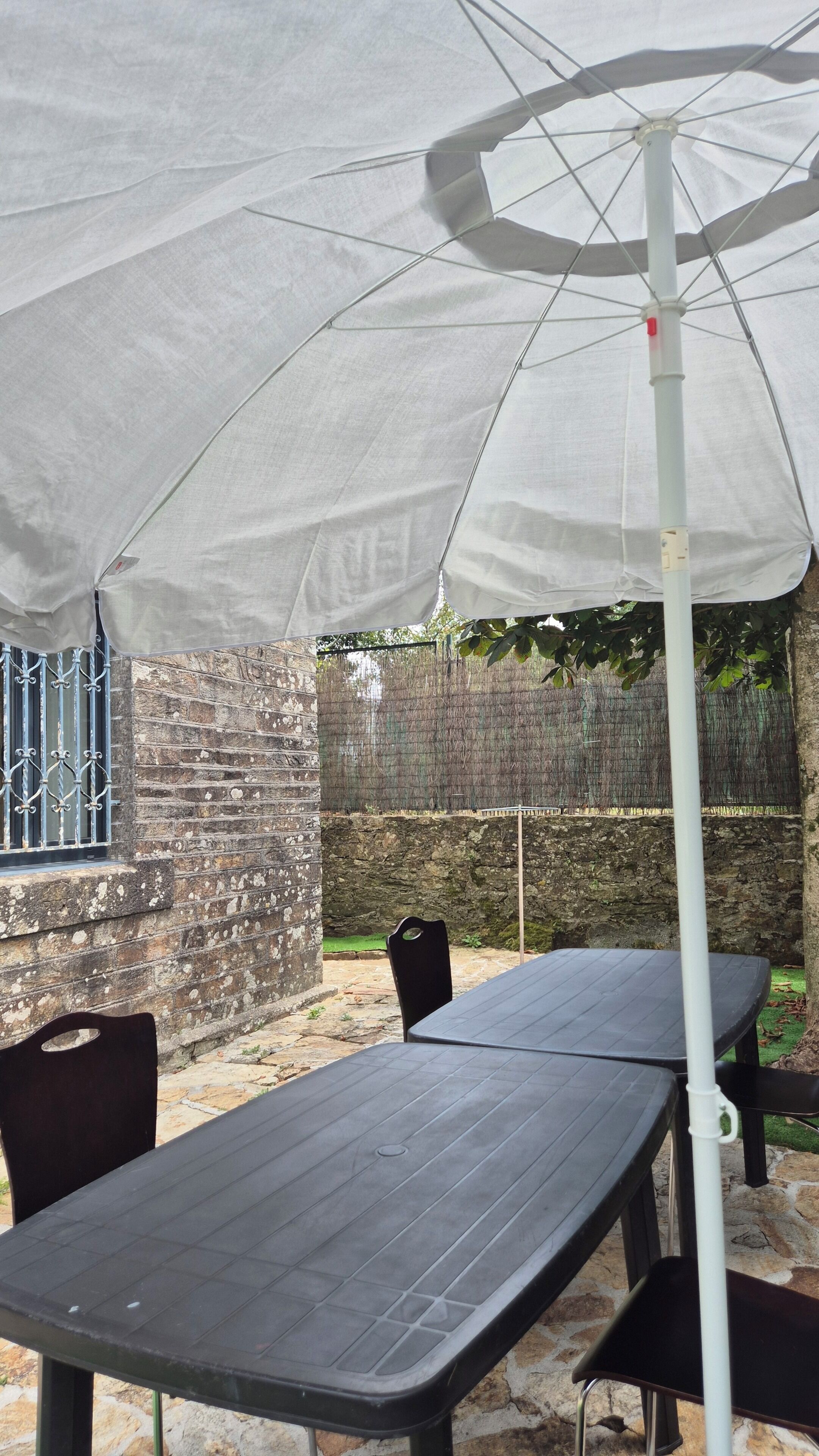 Un patio al aire libre con mesa y sillas, una sombrilla blanca y un muro de piedra al fondo.