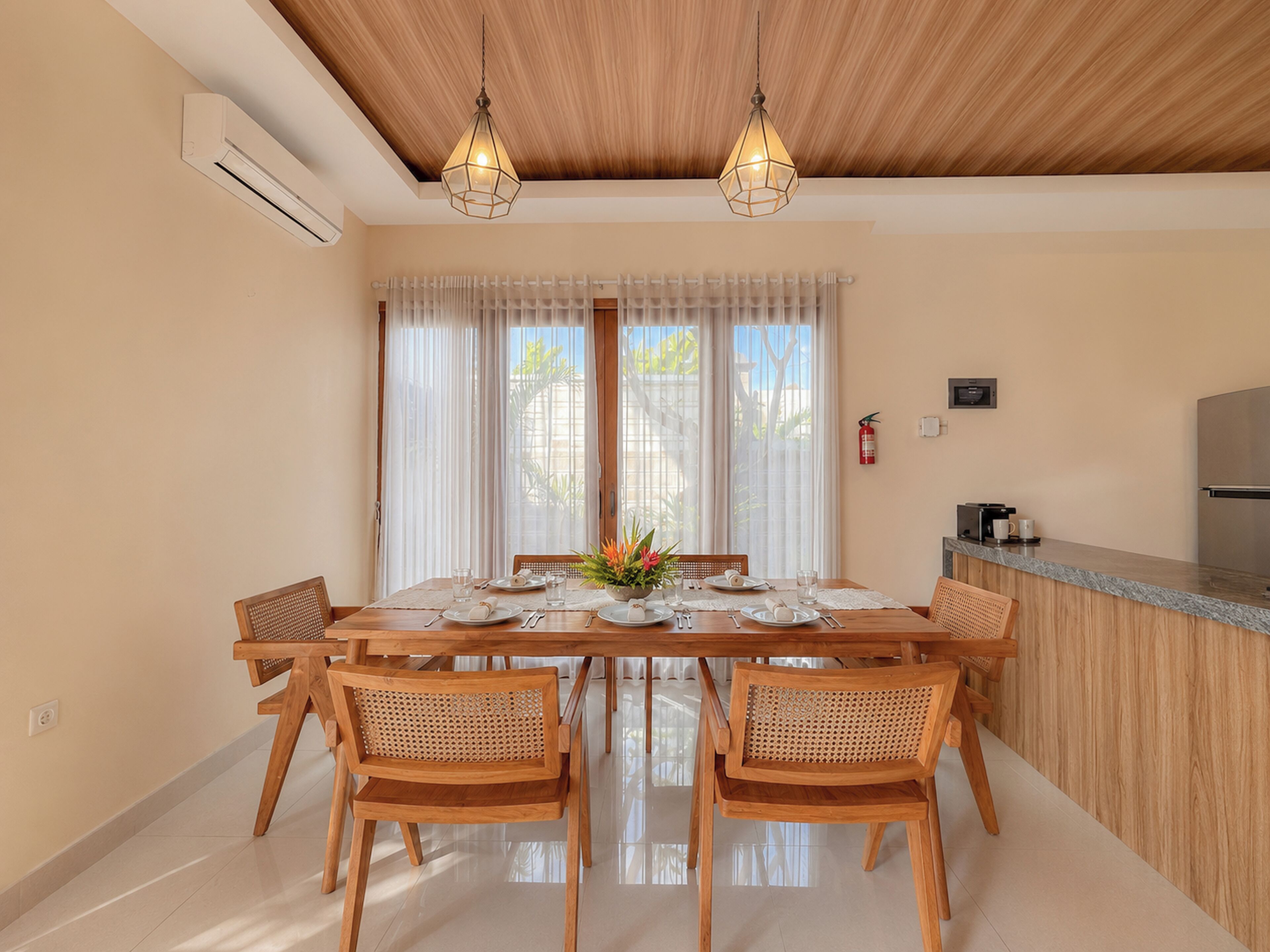 Comedor con mesa de madera dispuesta para cuatro personas, rodeada de sillas de mimbre, bajo un techo de madera con lámparas colgantes.