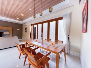 A dining area with a wooden table and chairs, large windows with curtains, and a ceiling with wooden panels and pendant lights. - Villa Amaya Canggu by Nagisa Bali (Badung)