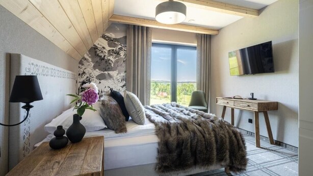 Ein Schlafzimmer mit einem Bett, einem Holzkopfende, einem Nachttisch mit Lampe, einem an der Wand befestigten Fernseher und einem Fenster mit Vorhängen.
