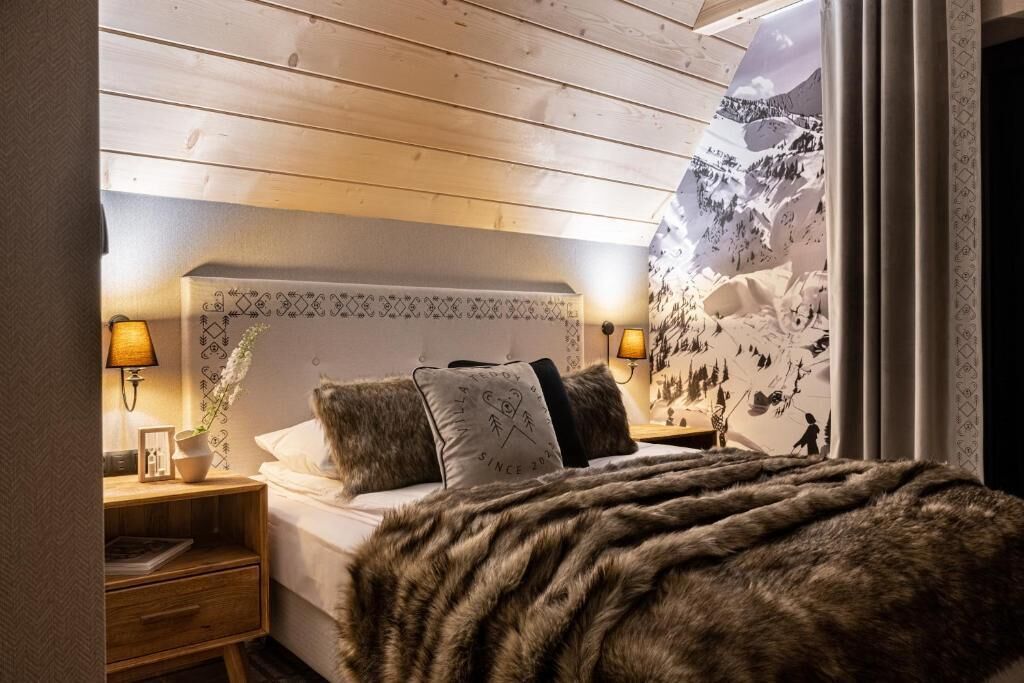 A bedroom with a wooden ceiling, a bed with a fur throw, two bedside lamps, and a wall with a mountain landscape.