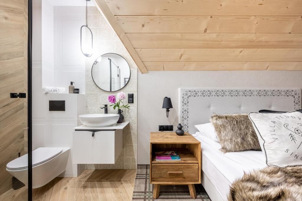 Un bagno moderno con lavandino, specchio e water bianchi. C'è un comodino con una lampada e un libro. La stanza ha un pavimento in legno e un letto bianco con un cuscino soffice.