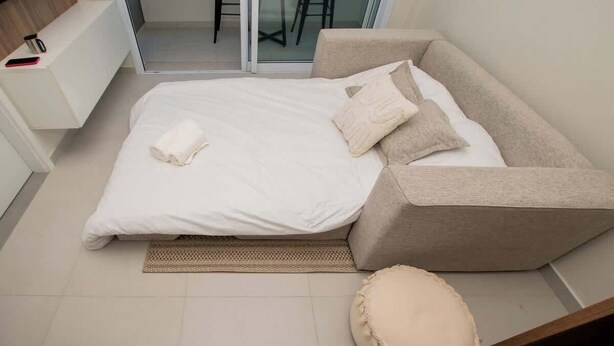 A bedroom with a bed, a sofa bed, a small round ottoman, and a white bedside table with a phone. - Corporate Stays First Living - 505 (Asunción)