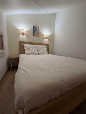 A bedroom with a wooden bed frame, two bedside lamps, a small bedside table, and a framed picture on the wall. - Le Petit Venard | Private parking | Private terrace (Vieux-Boucau-les-Bains)