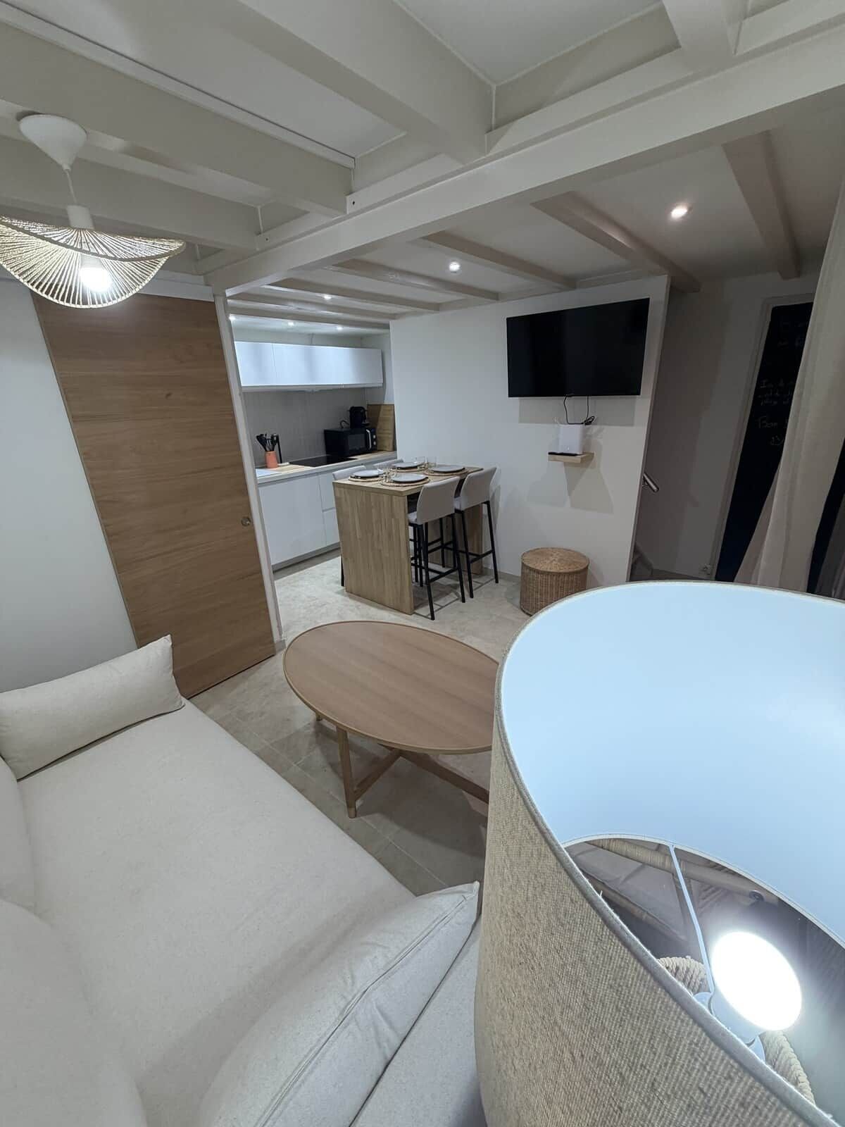 A modern hotel room with a sofa, a dining table, a kitchen area, and a TV mounted on the wall.