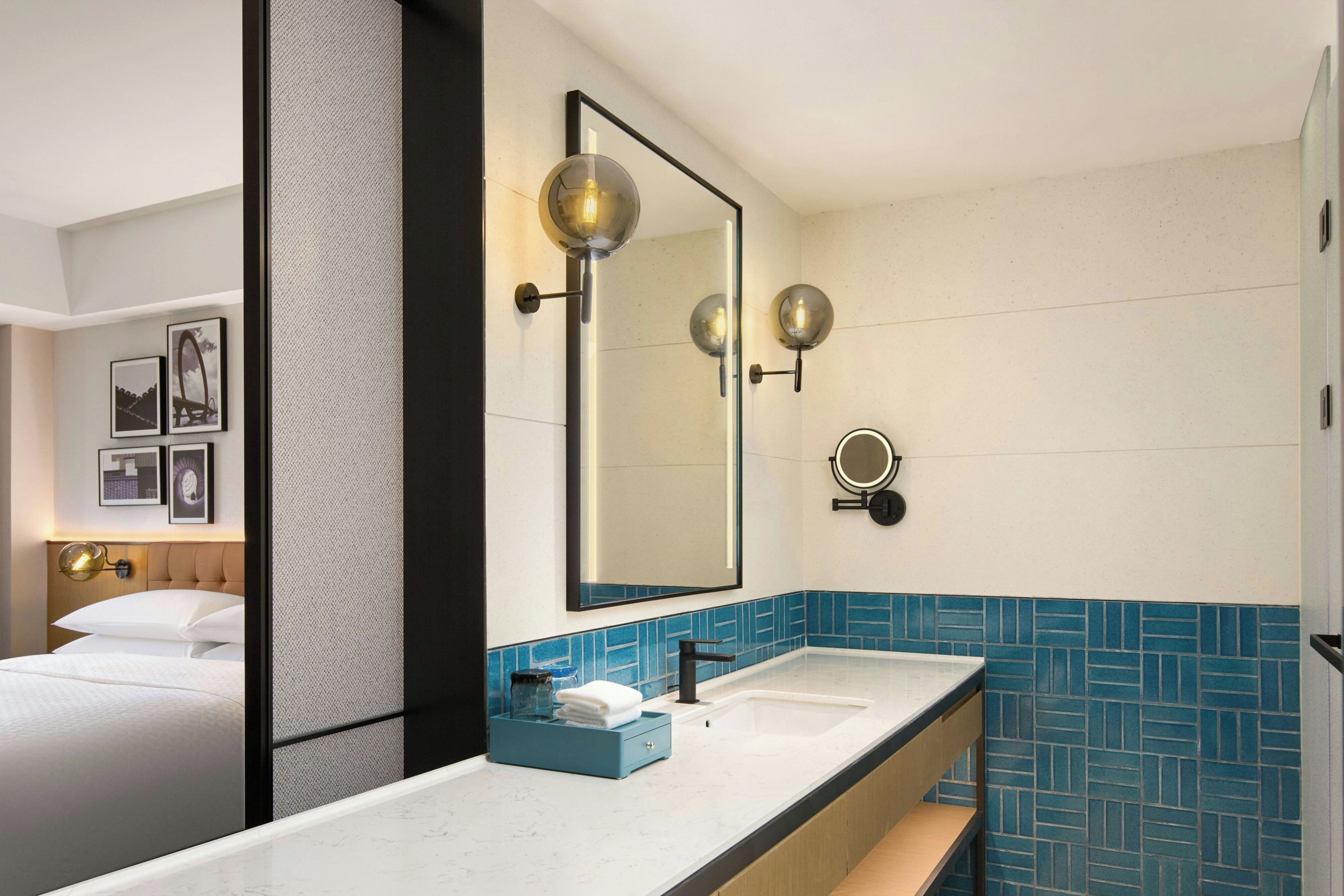 A hotel bathroom with a large mirror, two sinks, a blue-tiled wall, and a bed with white linens.