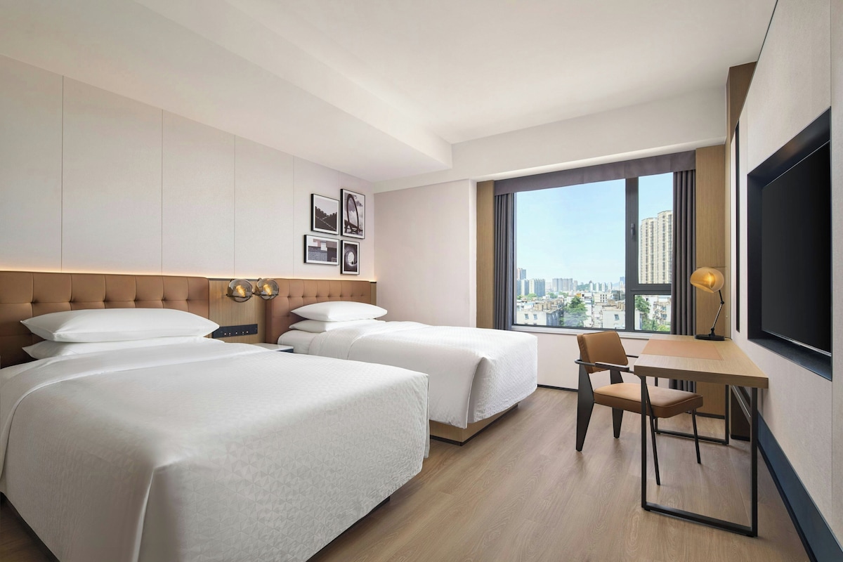 Habitación de hotel con dos camas, un escritorio, una silla, un televisor y un ventanal con vistas a la ciudad.