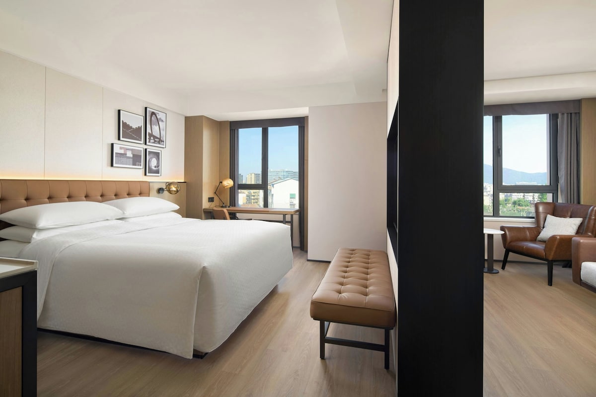 Habitación de hotel con una cama grande, un escritorio, un banco y vistas a la ciudad a través de las ventanas.
