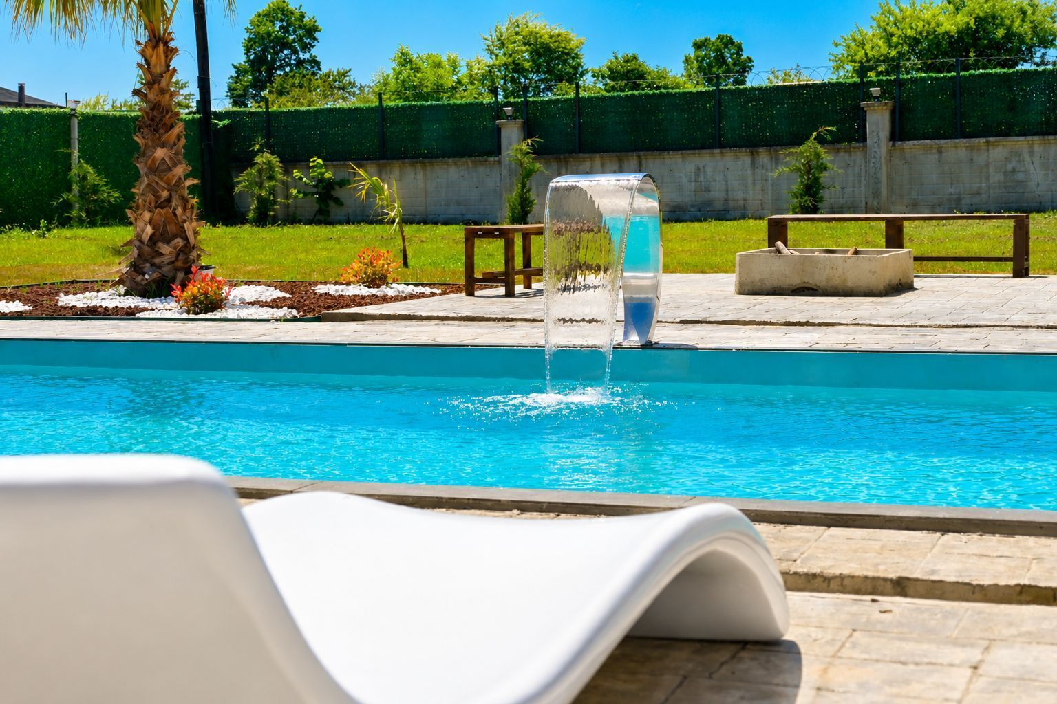 A swimming pool with a water feature, surrounded by a garden with a palm tree and benches.