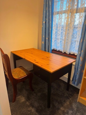 A corner room with a wooden table and a chair, a window with blue curtains, and a carpeted floor. - Modern 4-Bed Reading Home | Fast Wi-Fi & Garden (Reading)