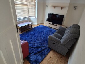 A living room with a blue rug, a grey sofa, a wooden coffee table, and a television on a stand. - Modern 4-Bed Reading Home | Fast Wi-Fi & Garden (Reading)