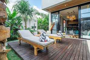 A modern outdoor lounge area with wooden deck, two lounge chairs, and a glass door leading to an interior space. - Tropical Blis Villa (Badung Regency)