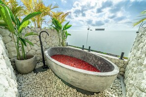 Eine Betonbadewanne, gefüllt mit roten Kieselsteinen, umgeben von weißen Kieselsteinen, Topfpflanzen und einer Steinmauer mit Meerblick.