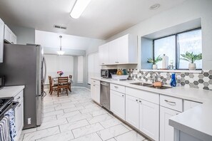 A modern kitchen with white cabinets, a stainless steel refrigerator, a dining area with a table and chairs, and a window with plants.