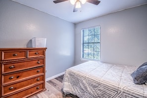 A bedroom with a wooden dresser, a bed with patterned bedding, a window with blinds, and a ceiling fan. - Deevine Homes - Private Bedroom with Shared Bathroom (2) (Carrollton)