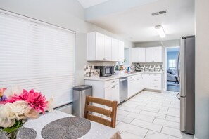 A modern kitchen with white cabinets, a dining table with flowers, and a view into the adjacent bedroom.
