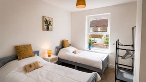 A bedroom with two beds, a bedside table with a lamp, a window with a view of buildings, and a clothes rack.
