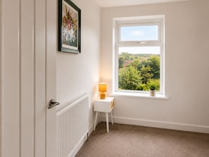A room with a white radiator, a small white table with a lamp, a framed picture on the wall, and a window with a view of greenery.