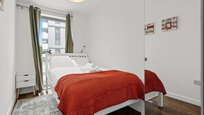 A bedroom with a bed, a bedside table, a lamp, a window with curtains, and artwork on the wall. - Modern 2-Bed Flat with Balcony near Canary Wharf (London)