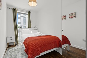Ein Schlafzimmer mit einem Bett, einem Nachttisch, einer Lampe, einem Fenster mit Vorhängen und einem Bild an der Wand.