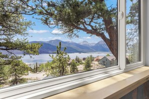 Blick durch ein Fenster auf ein Seeufergrundstück mit Bergen im Hintergrund.