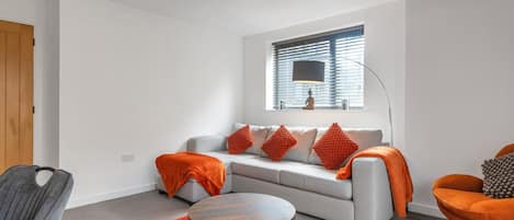 A modern living room with a grey sofa, orange throw pillows, a round coffee table, and a window with blinds.