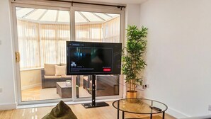 A modern living room with a flat-screen TV on a stand, a glass coffee table, a sofa with a green pillow, and a potted plant. - Modern 2-Bed House Near Central Canals & Arena (Birmingham)