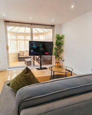 A modern living room with a flat-screen TV on a stand, a glass coffee table, a sofa with a green pillow, and a potted plant. - Modern 2-Bed House Near Central Canals & Arena (Birmingham)