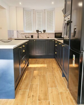 A kitchen with dark cabinets, a stainless steel refrigerator, and a window with blinds. - Modern 2-Bed House Near Central Canals & Arena (Birmingham)