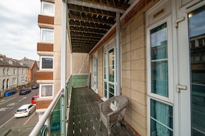 Un balcon avec un fauteuil en osier, une vue sur une rue bordée de voitures garées et un immeuble doté de nombreuses fenêtres.