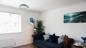 Una sala de estar con un sofá azul oscuro, una lámpara de pie, una planta en maceta y un cuadro de una playa.