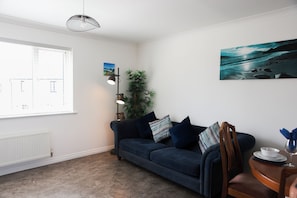 Ein Wohnzimmer mit einem dunkelblauen Sofa, einer Stehlampe, einem Blumentopf und einem Gemälde mit einer Strandansicht.
